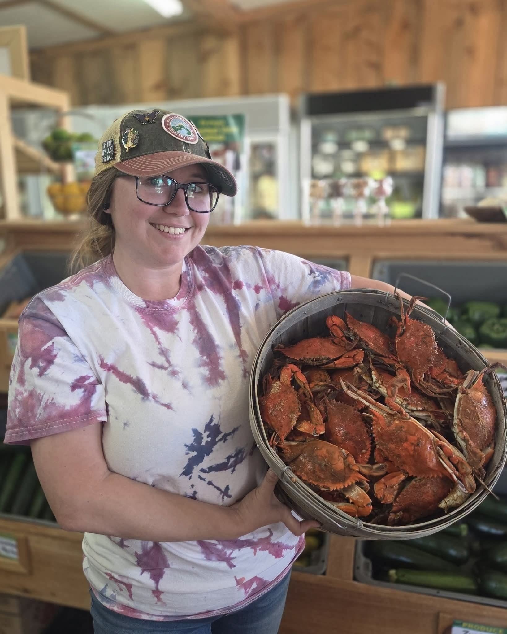 Smiling woman in a tie-dye shirt and cap holding a basket full of cooked crabs indoors.