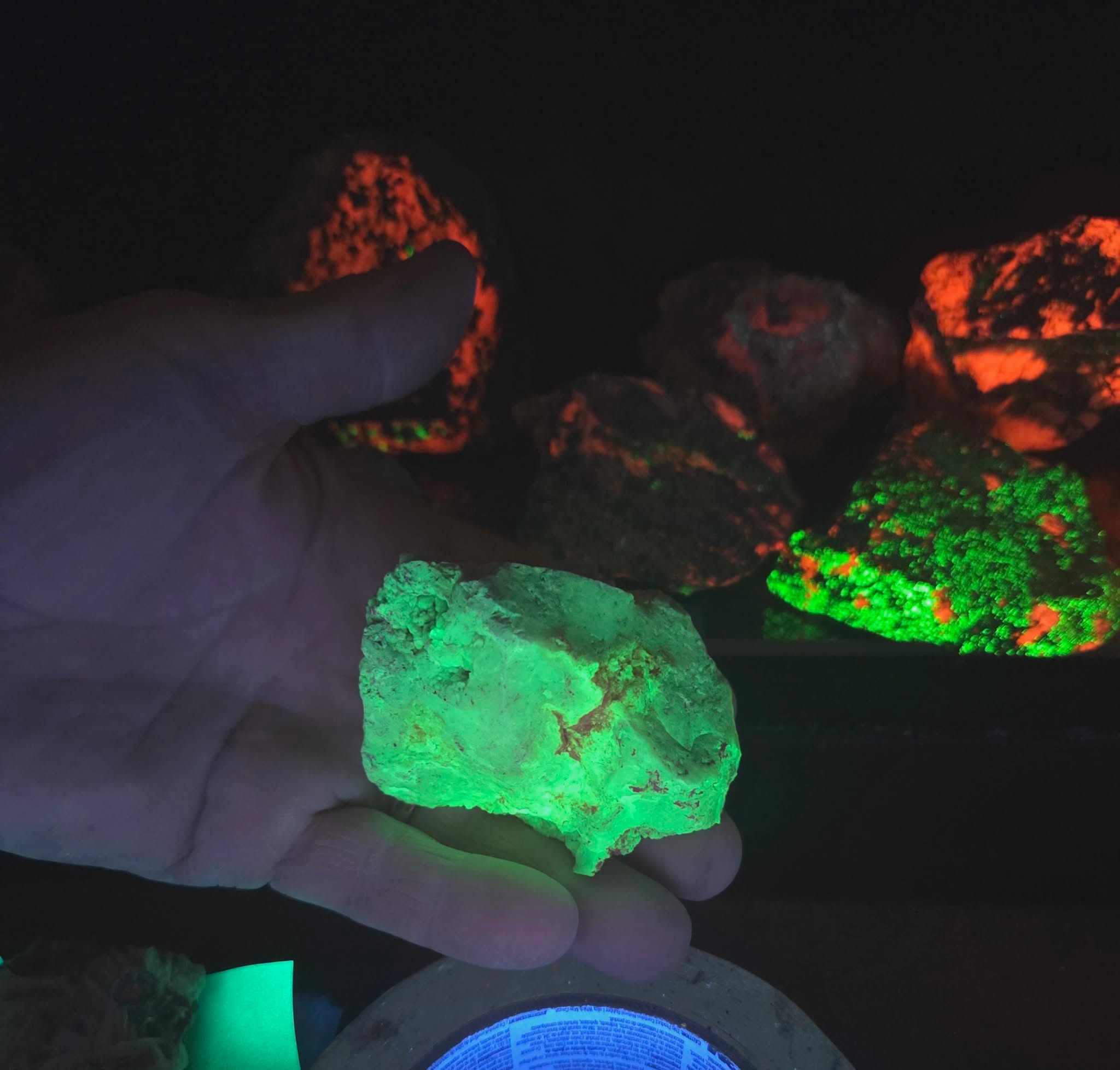Hand holding a glowing green fluorescent mineral rock with other glowing rocks in the dark background.