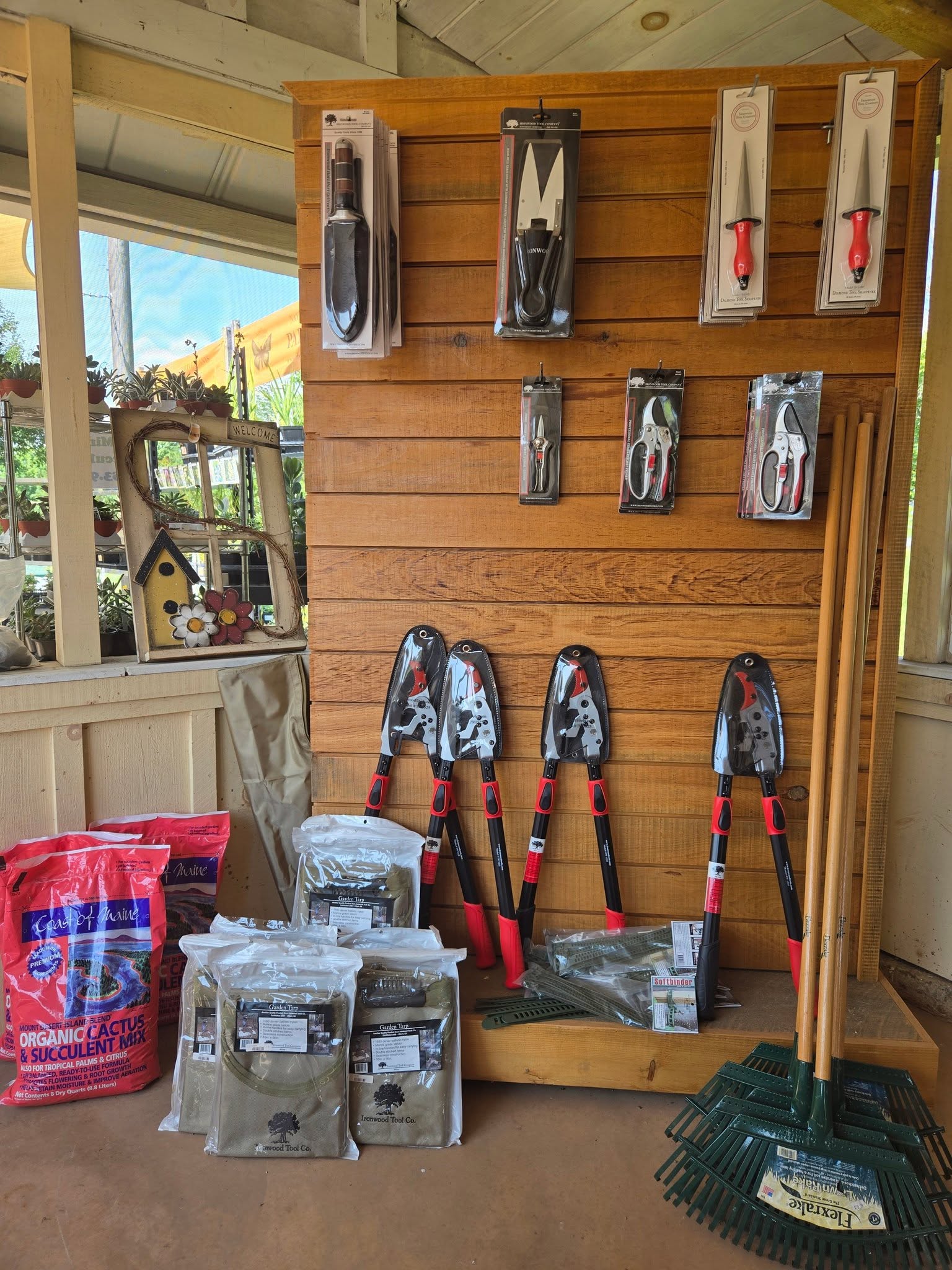 Wooden wall display with gardening tools, rakes, organic cactus soil, and protective gear in a garden shop.