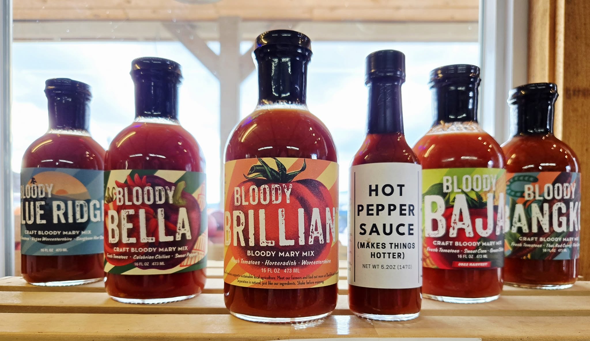 Six Bloody Mary mix and hot pepper sauce bottles lined up on a wooden shelf by a window.