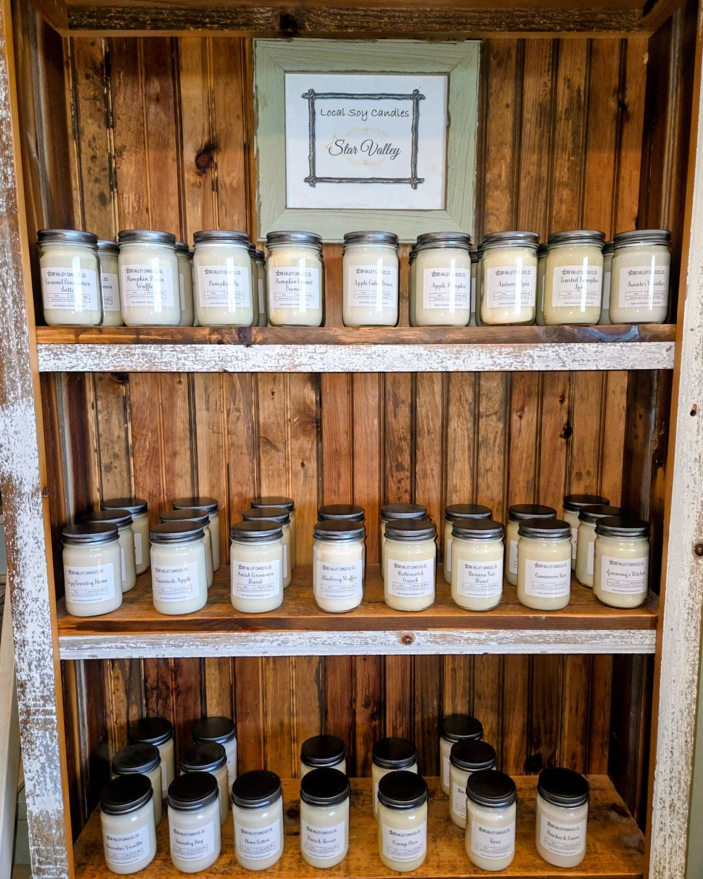 Wooden shelves with three rows of labeled soy candles in glass jars with metal lids.