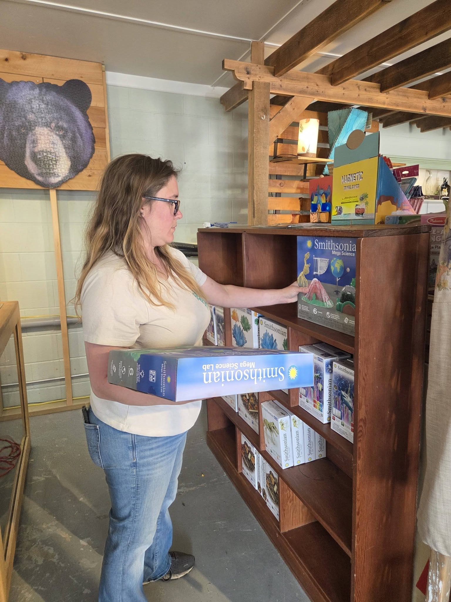 Woman with glasses holding a Smithsonian Mega Science Lab box while selecting another from a wooden shelf.