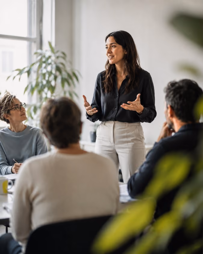 A woman standing and speaking to three seated colleagues in a bright office meeting room.
