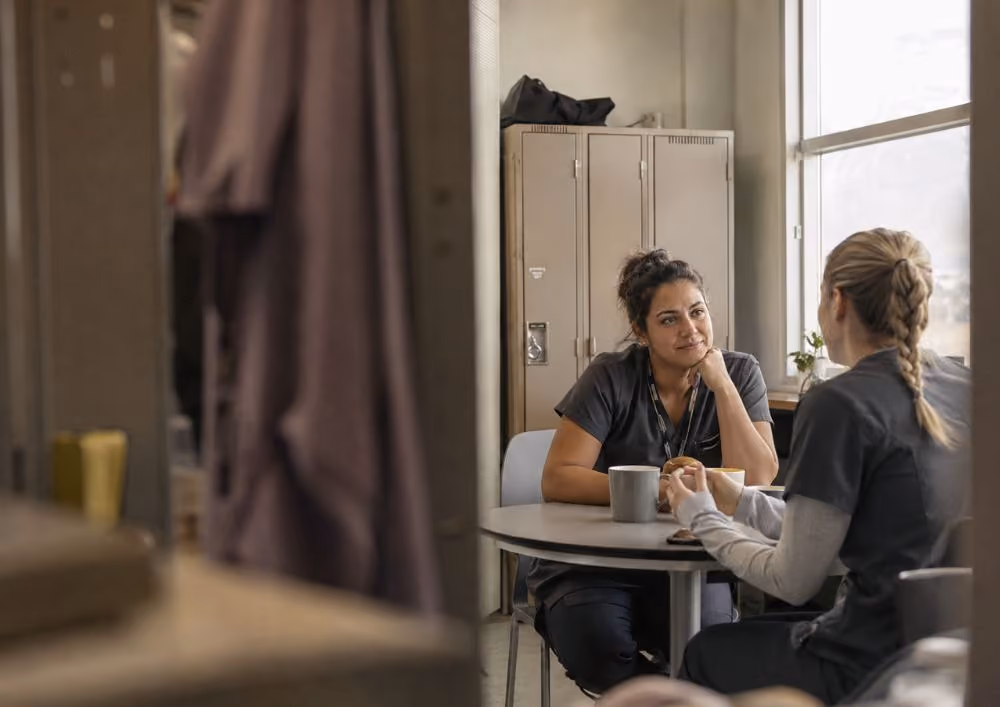 Two healthcare workers in scrubs talking over coffee at a round table in a break room.
