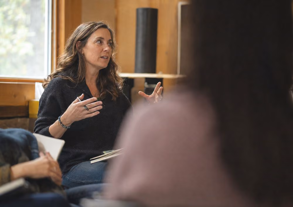 Woman speaking and gesturing during a group discussion in a cozy room with wooden walls and window.