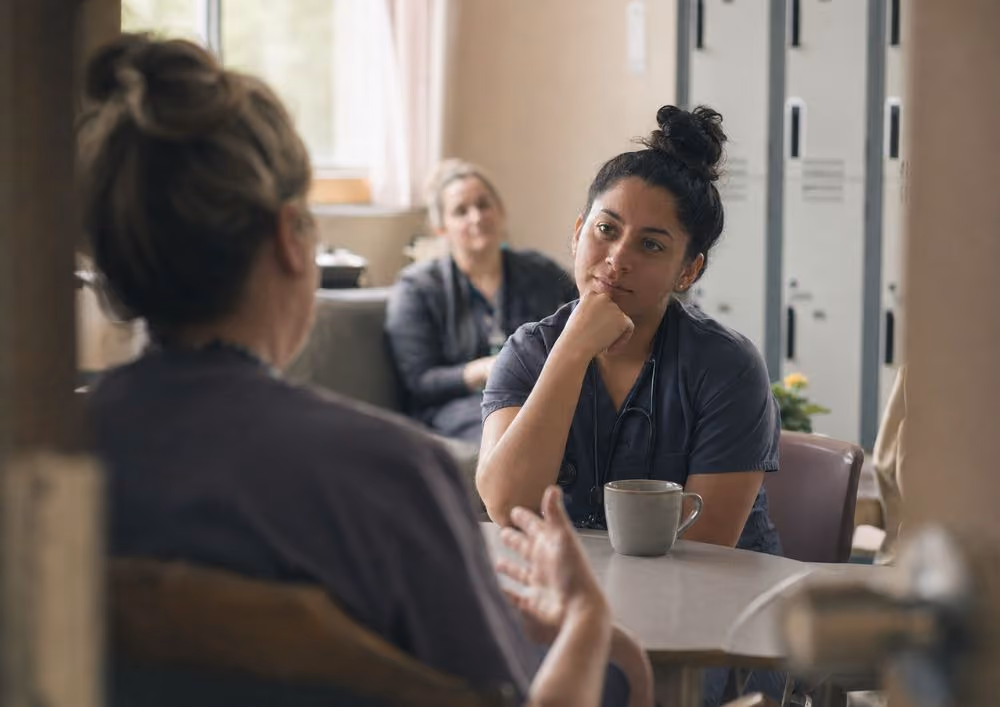 Healthcare worker in scrubs attentively listening to a colleague at a table with a mug.