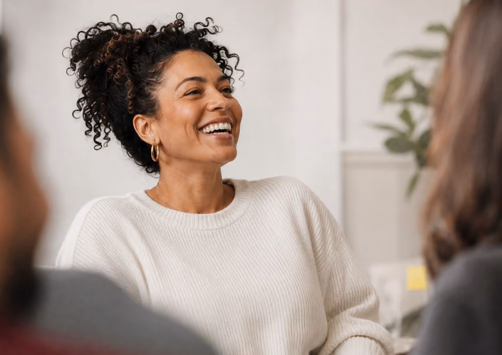 Smiling woman with curly hair and gold hoop earrings wearing a cream sweater in a group setting.