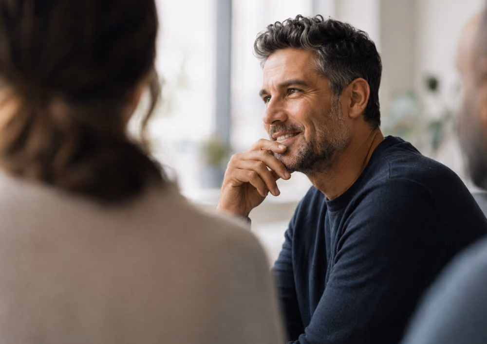 Smiling man listening thoughtfully to two people in a bright room with a window in background.