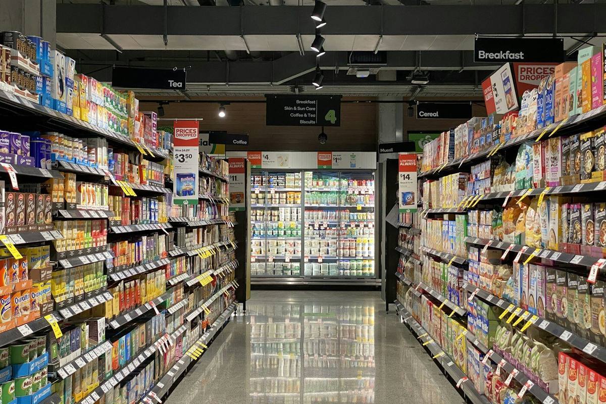 Empty supermarket aisle with rows of shelving, track lighting on and refrigerated display cases in the background