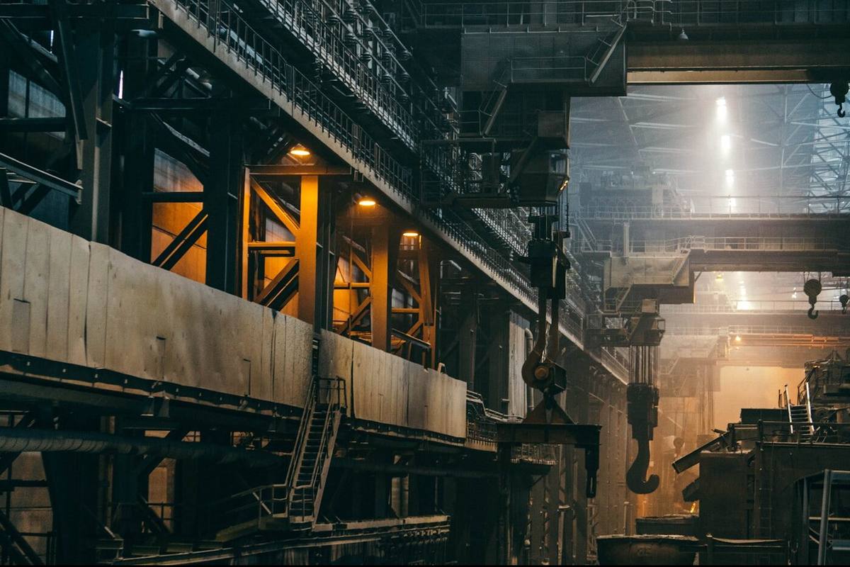 Interior of a heavy industrial hall with an overhead crane, steel structures and the glow of a hot process in the background