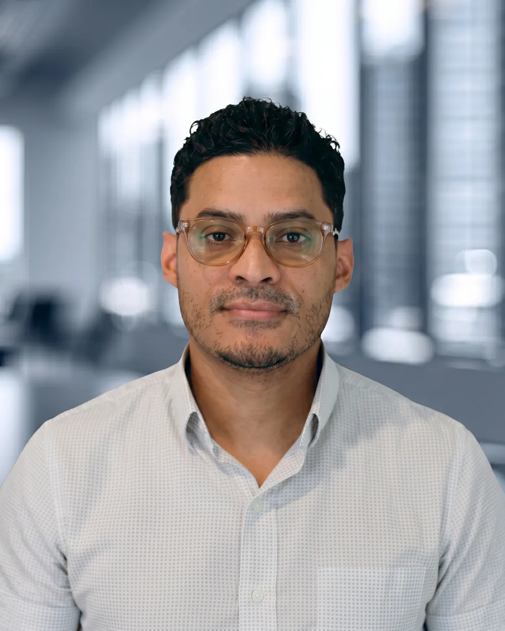 Man wearing glasses and a light patterned shirt in a bright, blurred office background.