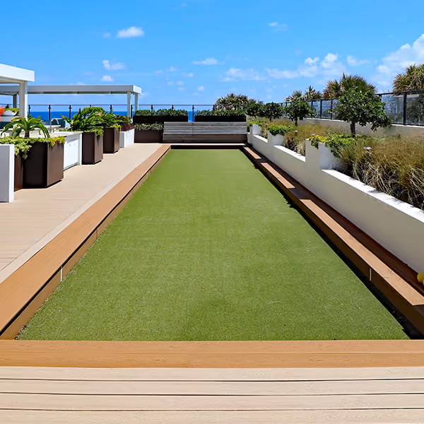 Outdoor bocce ball court with artificial turf surrounded by plants and ocean view under blue sky.