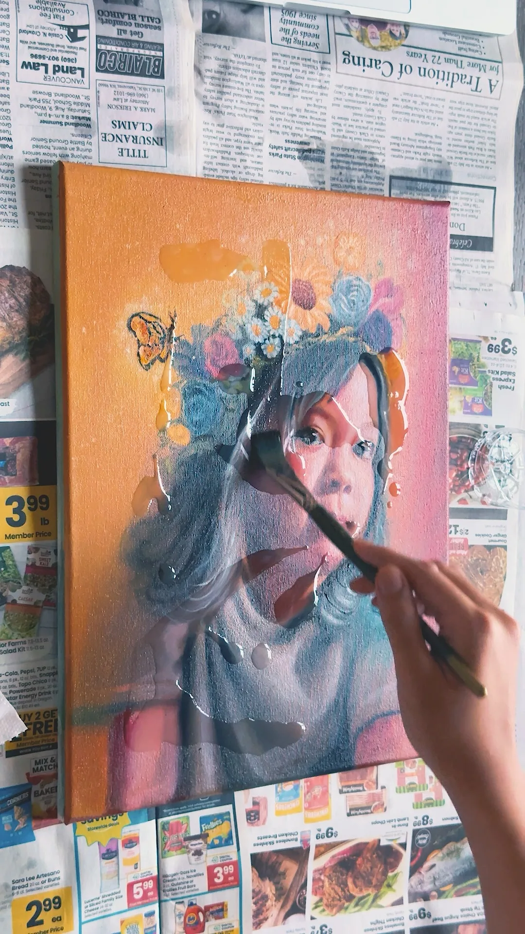 Still from the varnishing video, the flower crown painting lying on newspaper while a brush spreads varnish across the wet surface.