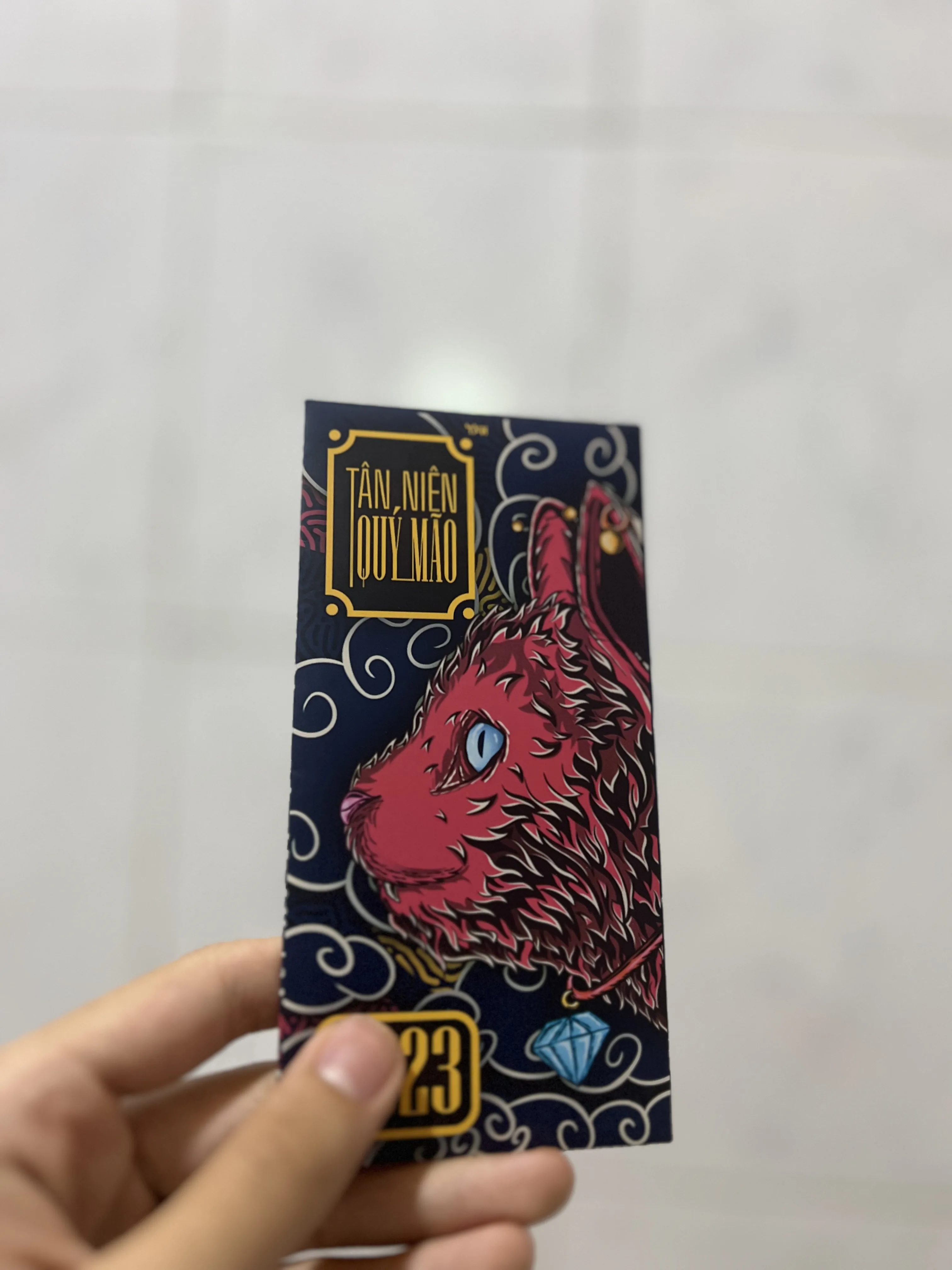 Hand holding the printed red envelope, navy with gold cloud patterning, a magenta cat profile and a cyan gem, lettered Tân Niên Quý Mão and 2023.