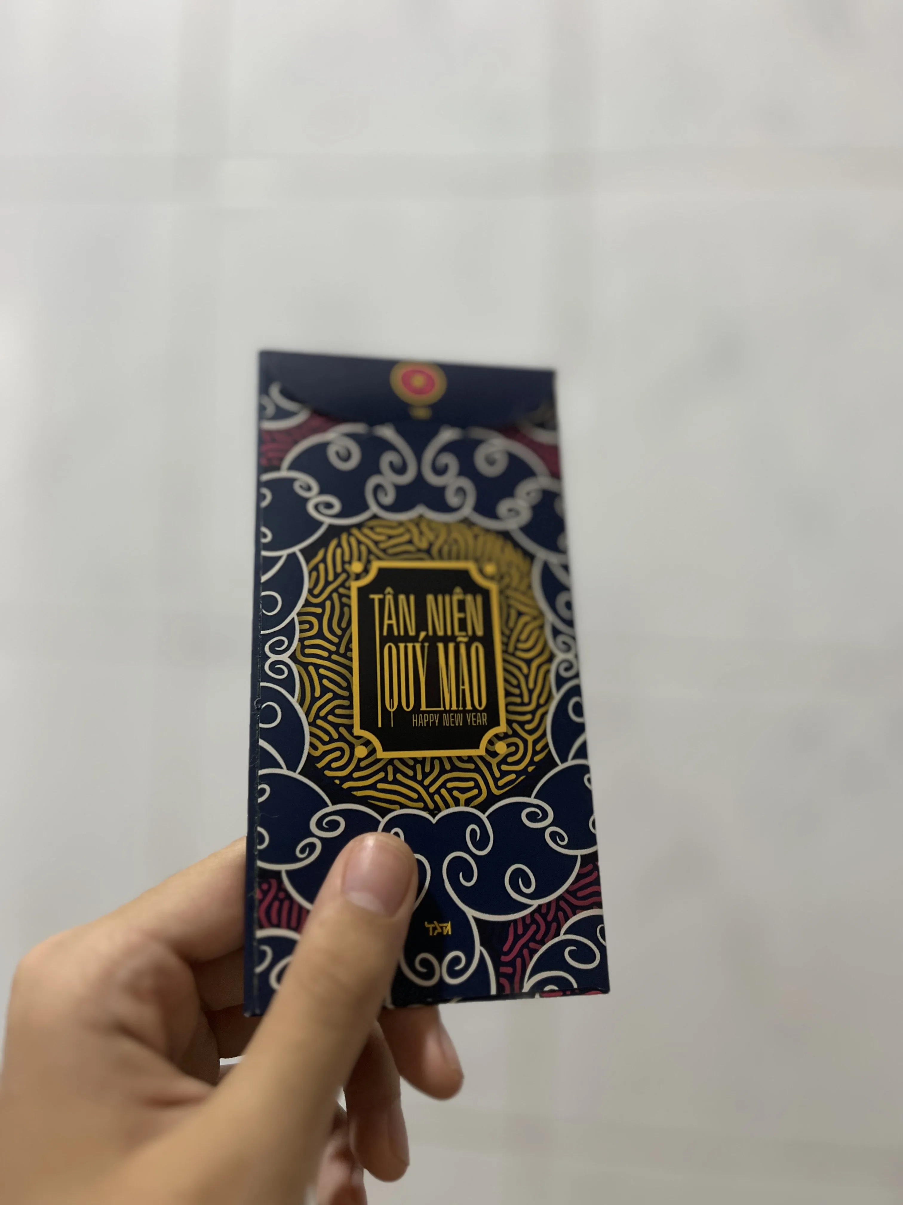 Back of the printed red envelope held in a hand, a gold medallion set in navy and red cloud patterning, lettered Tân Niên Quý Mão and Happy New Year.