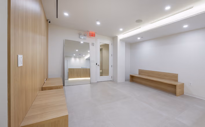 Minimalist waiting room with wooden benches, white walls, and an exit sign above a white door.