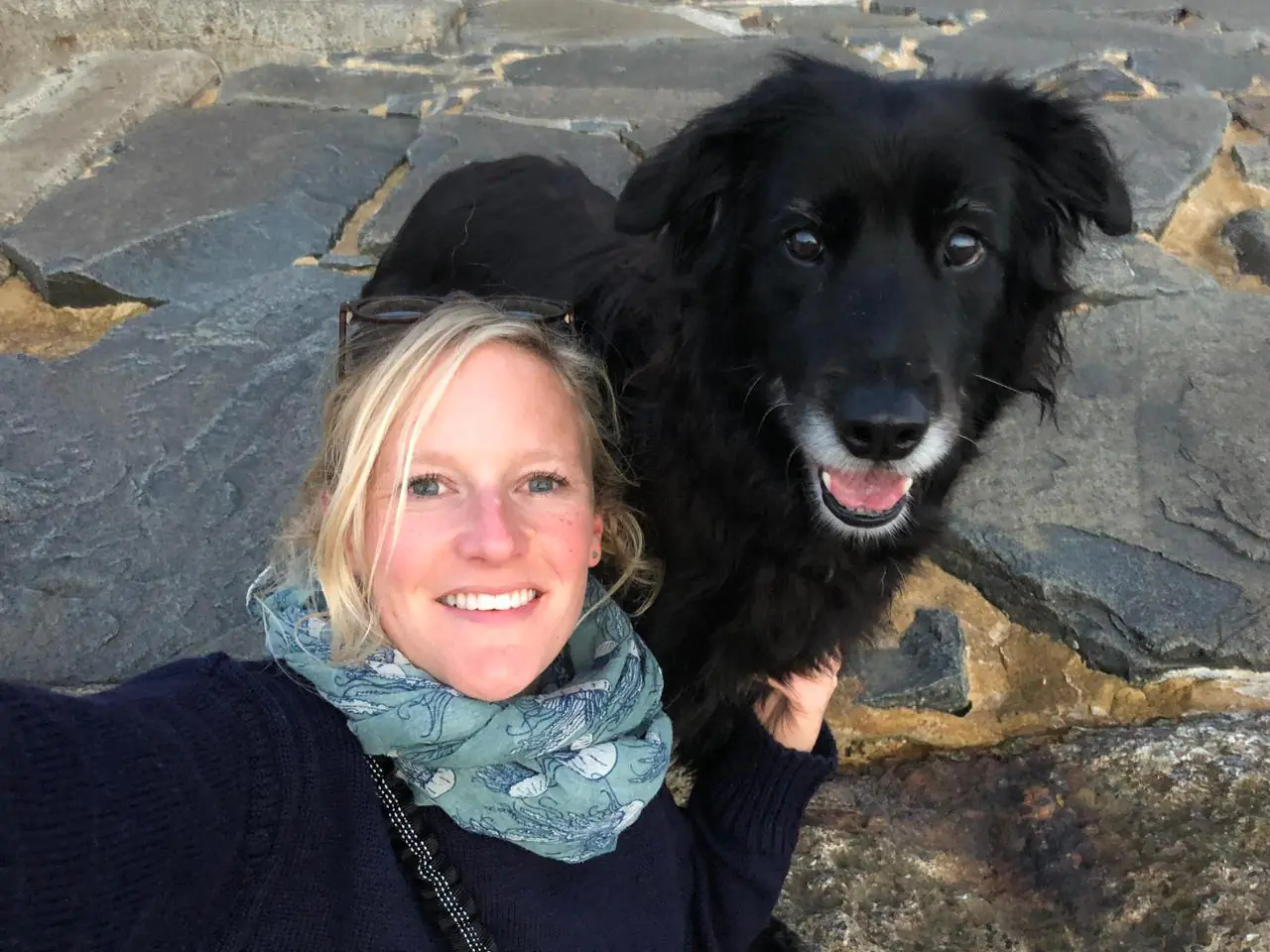 Sarah wearing a scarf taking a selfie with a happy black dog on stone pavement.