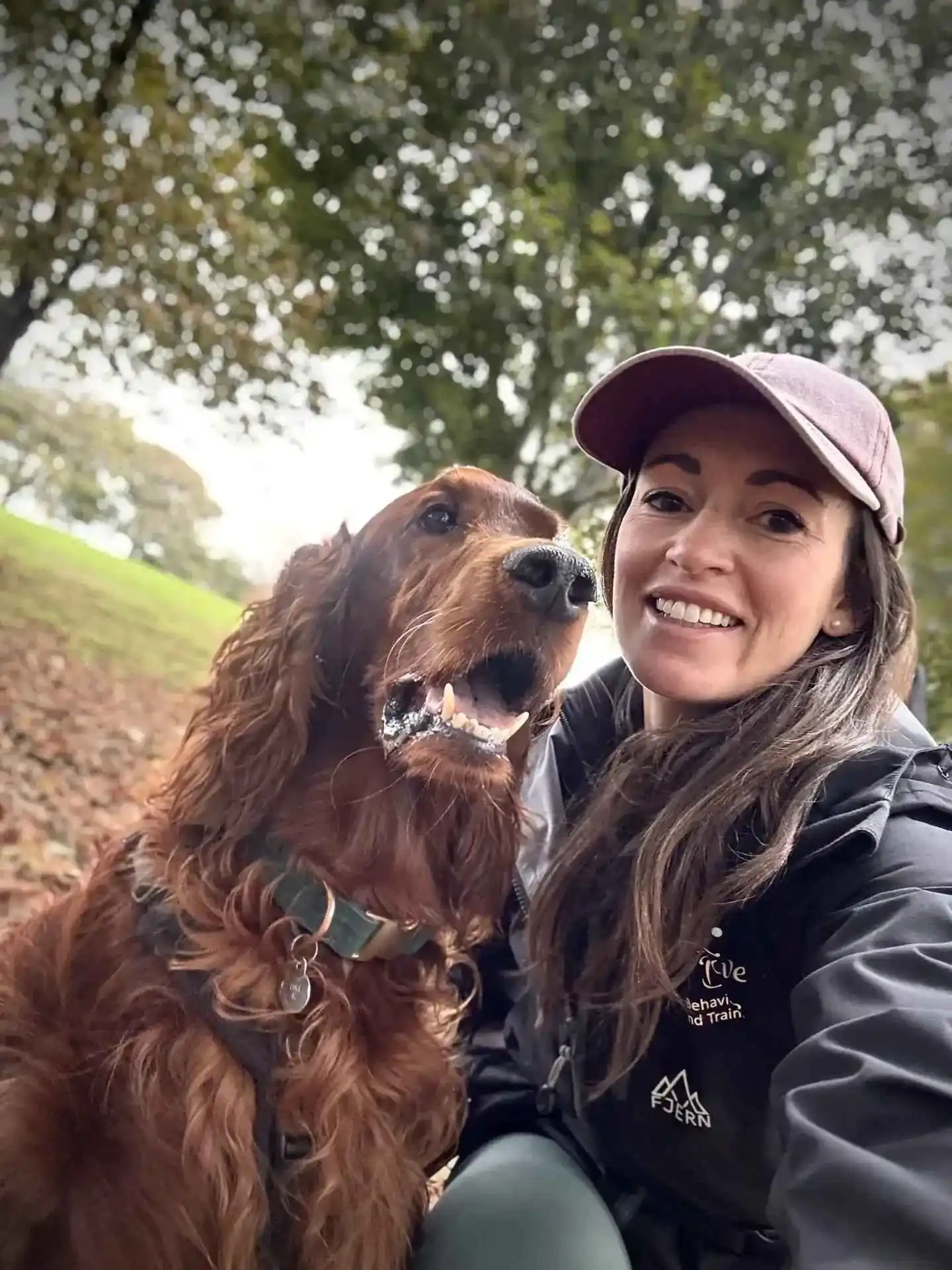 Katie wearing a cap smiling with a happy brown Irish Setter dog outdoors with trees in background.