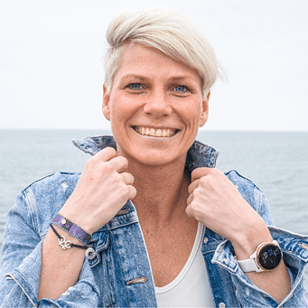 Lächelnde Frau mit kurzem blondem Haar trägt Jeansjacke und Armbanduhr, steht vor ruhiger Meereskulisse.