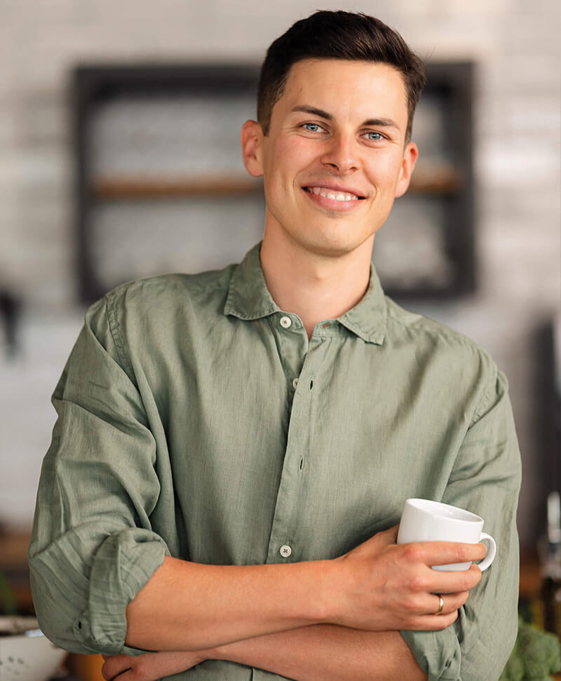 Junger lächelnder Mann mit kurzem dunklem Haar trägt ein grünes Hemd mit hochgekrempelten Ärmeln und hält eine weiße Tasse in der Hand vor verschwommenem Hintergrund.