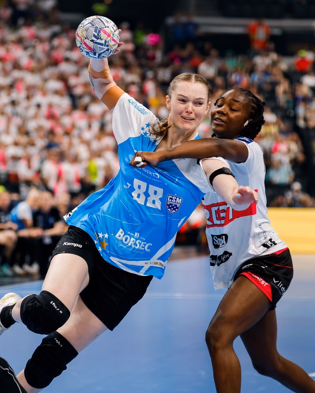 Handball player in blue tries to throw ball while opponent in white defends closely.