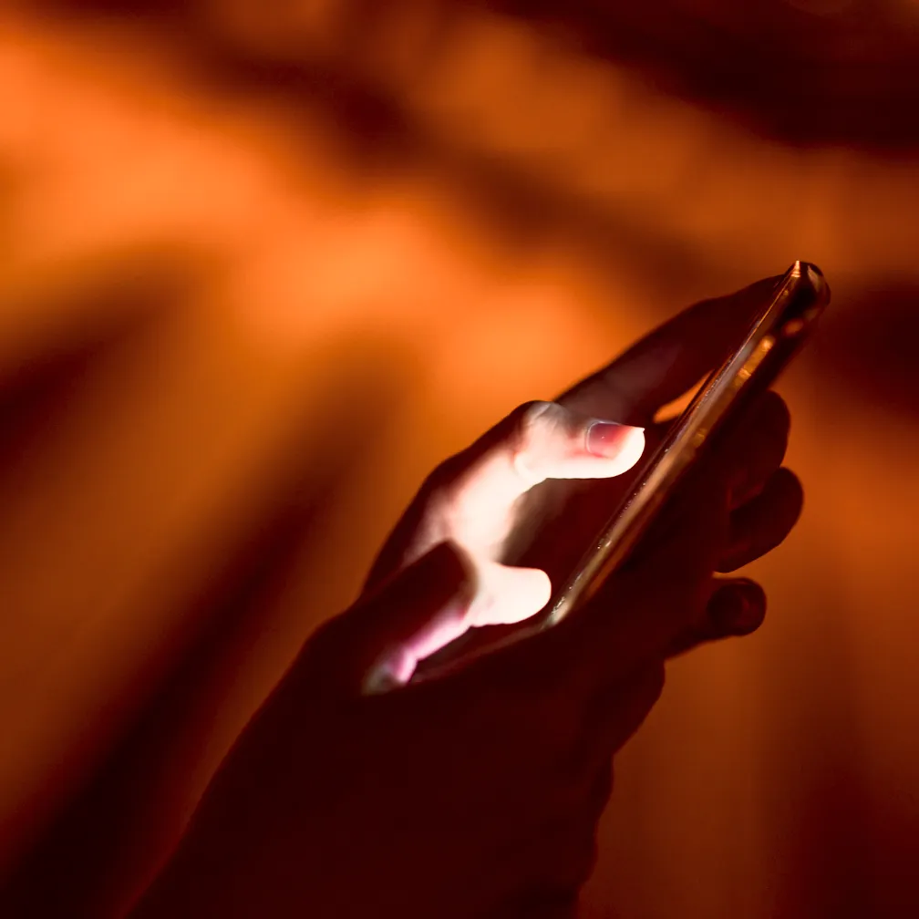 Close-up of hands holding and using a smartphone with the screen lighting the fingers against a blurred orange background.