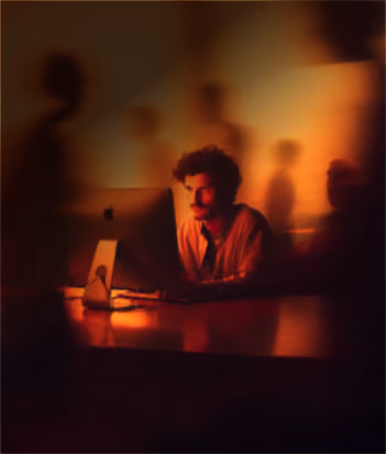 A man with curly hair sits at a desk working on a computer in a dimly lit room with blurred shadowy figures around him.