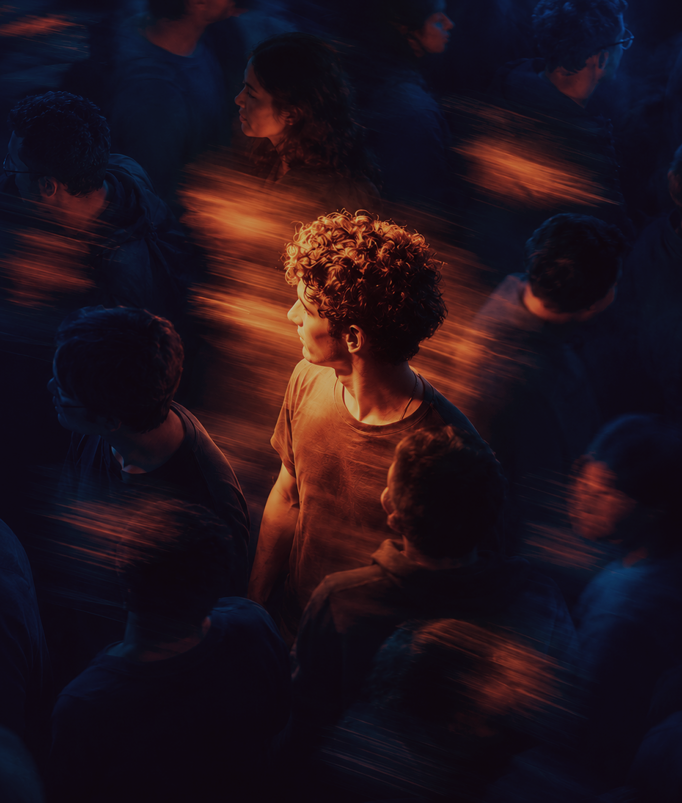 Young man with curly hair and orange lighting stands out facing left among blurred crowd in dark surroundings with streaks of orange light.