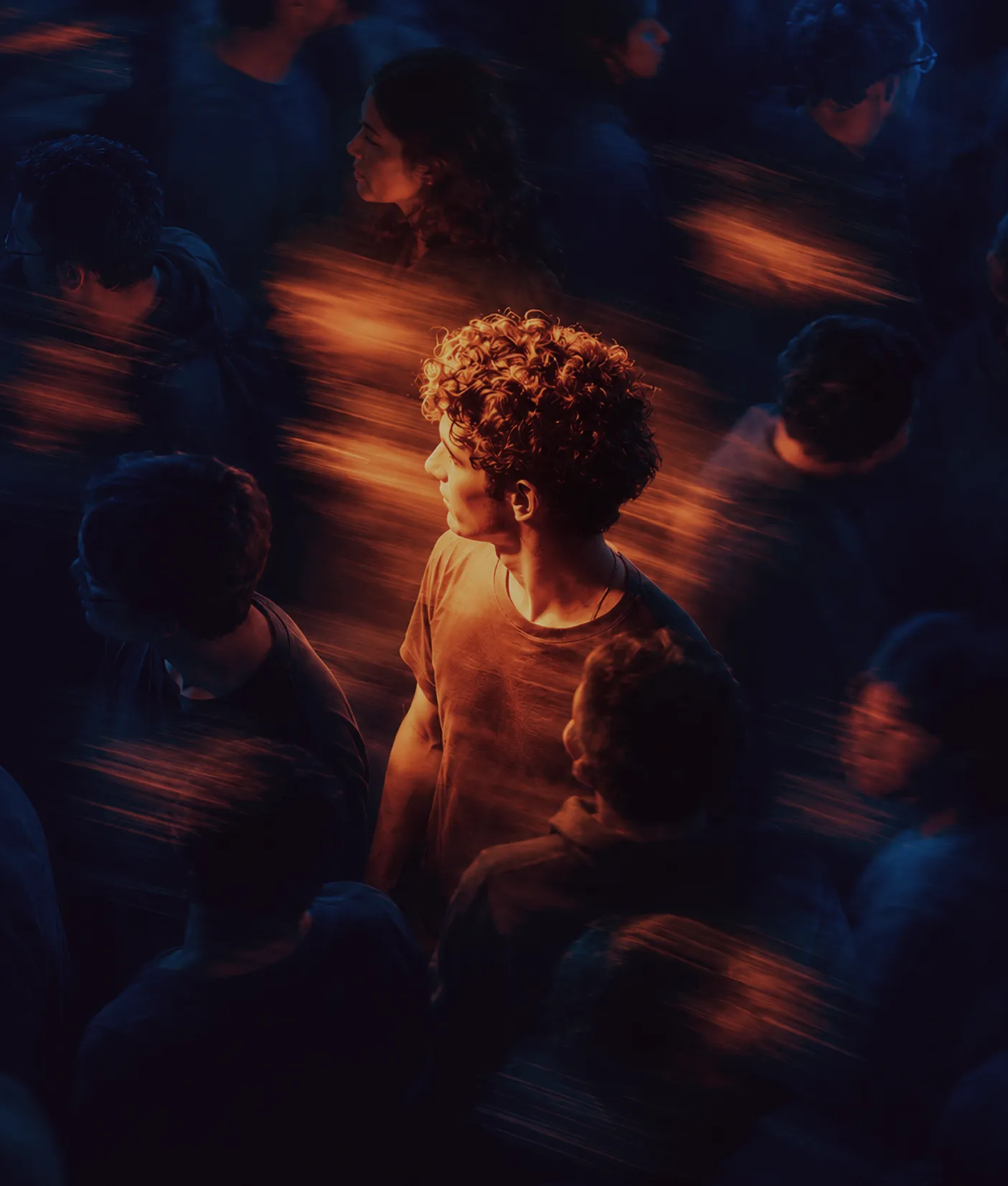 Young man with curly hair and orange lighting stands out facing left among blurred crowd in dark surroundings with streaks of orange light.