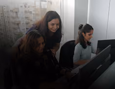Three women working together at computers in a dimly lit office, collaborating closely.