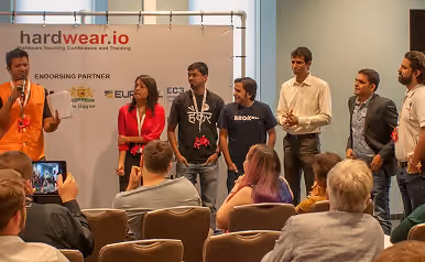 Seven people standing in a row on a stage during a presentation or panel discussion at a conference, with an audience seated in front of them. A banner behind them reads 'hardwear.io' and displays sponsor logos.