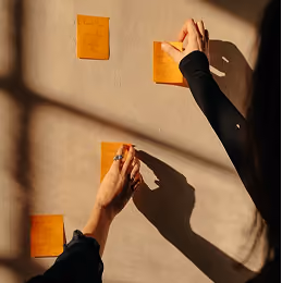Person placing or arranging four orange sticky notes on a beige wall with sunlight casting shadows.