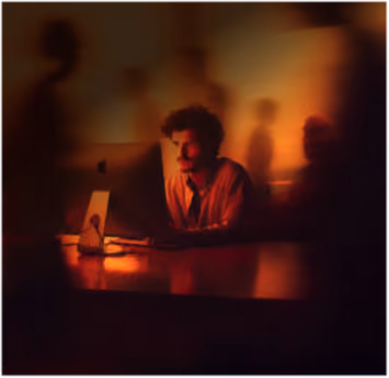 A dimly lit scene with a man sitting alone at a table illuminated by warm, orange light, surrounded by blurred shadows of other figures.