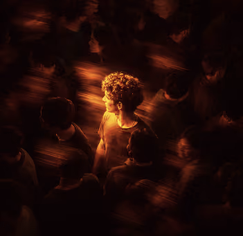 A person with curly hair illuminated by warm light stands out in a crowd of blurred figures in darkness.