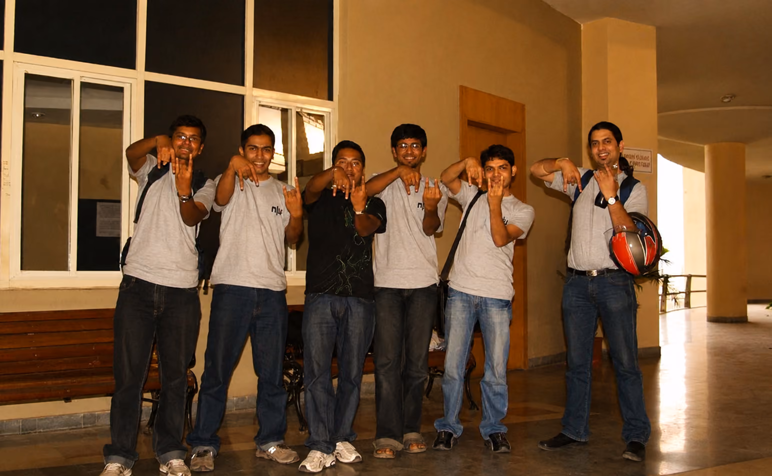 Group of six young men standing indoors in a hallway, five wearing gray t-shirts with backpacks, one in black shirt, all making hand gestures toward the camera and smiling.