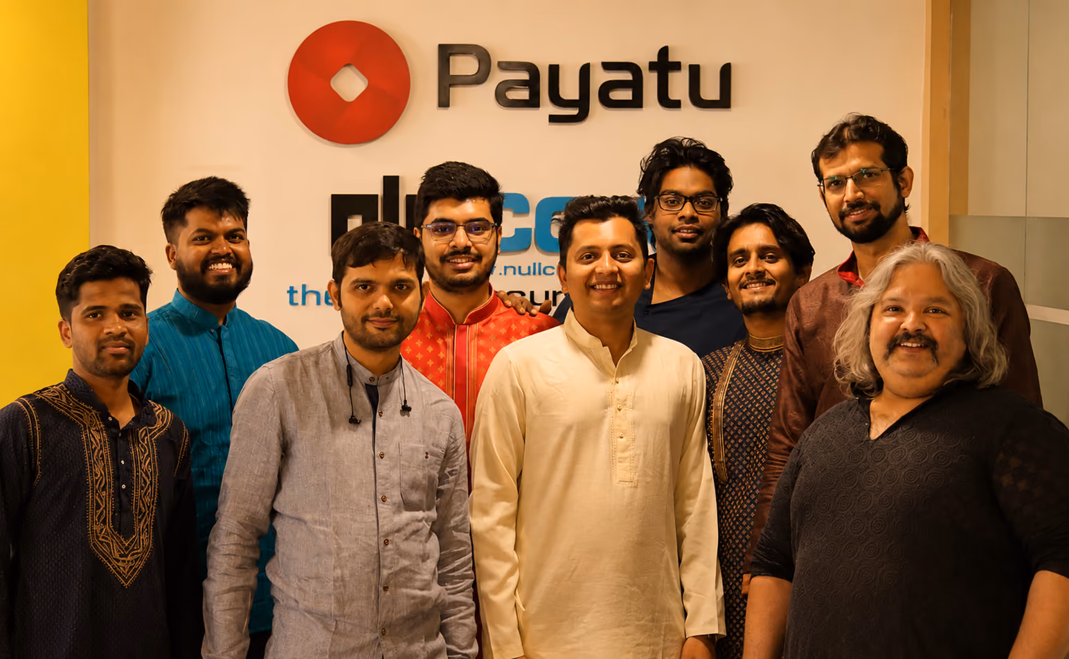 Group of nine men standing and smiling in front of a wall with Payatu and other company logos, all wearing casual or ethnic clothing.