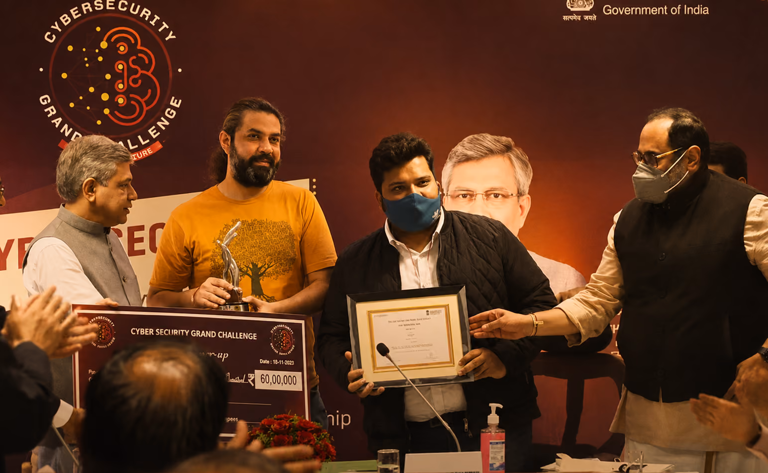 Group of men at an award ceremony for the Cyber Security Grand Challenge; one man holding a large check of 6,000,000 rupees, another holding a framed certificate, with a backdrop featuring the event logo and the Government of India emblem.