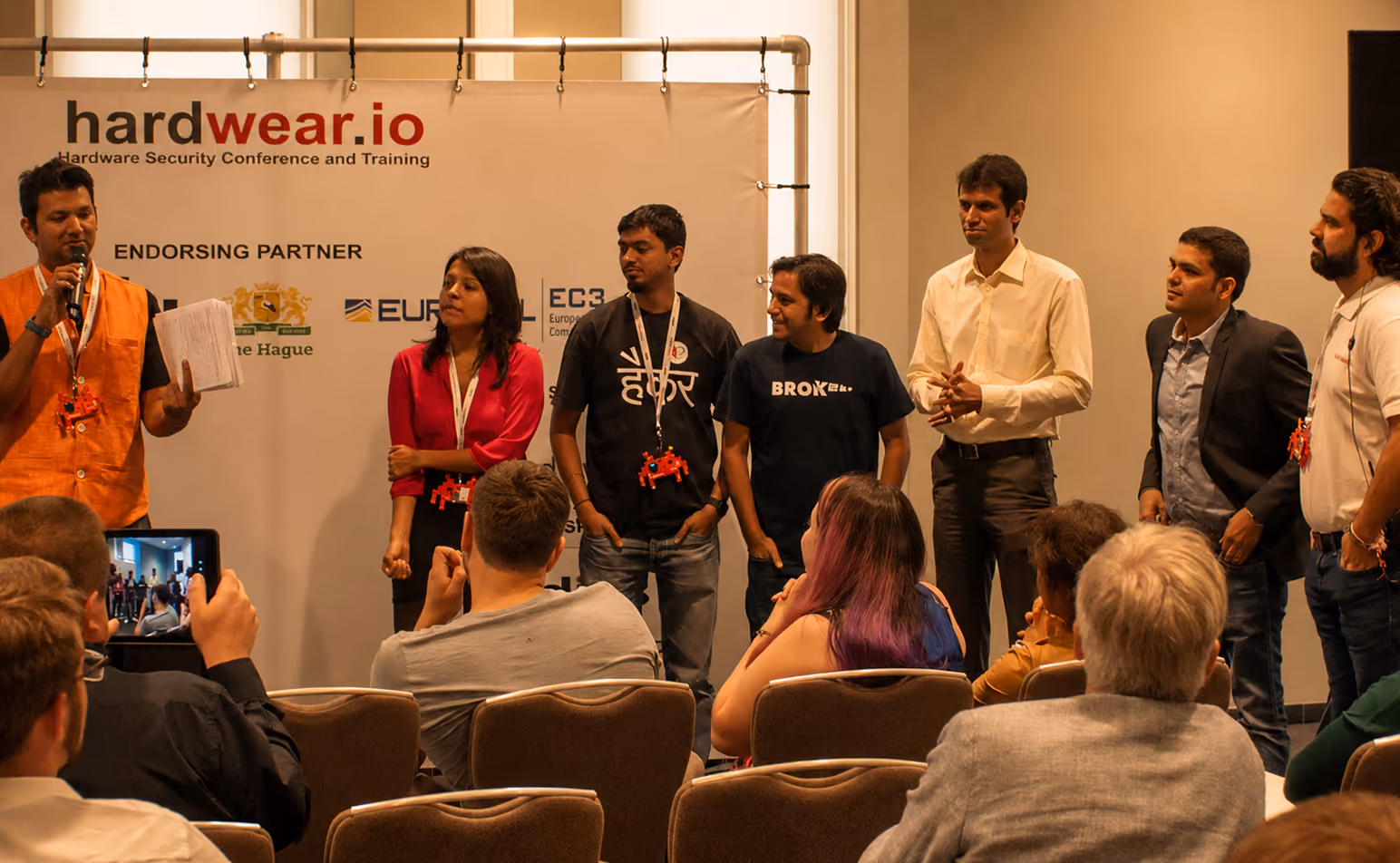 A group of seven people standing in front of a conference backdrop with the logo 'hardwear.io Hardware Security Conference and Training,' addressing an audience seated in chairs, one person speaking into a microphone while others listen. The audience includes individuals with varied hair colors, including one person with purple hair, engaged and watching the speakers.