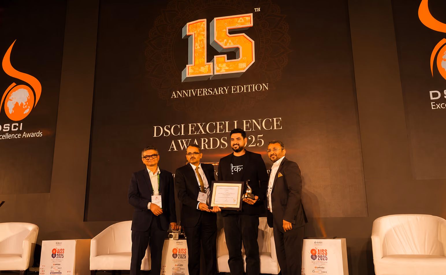 Four men standing on a stage at the 15th Anniversary Edition of the DSCI Excellence Awards 2025, with one man holding a certificate and a trophy.