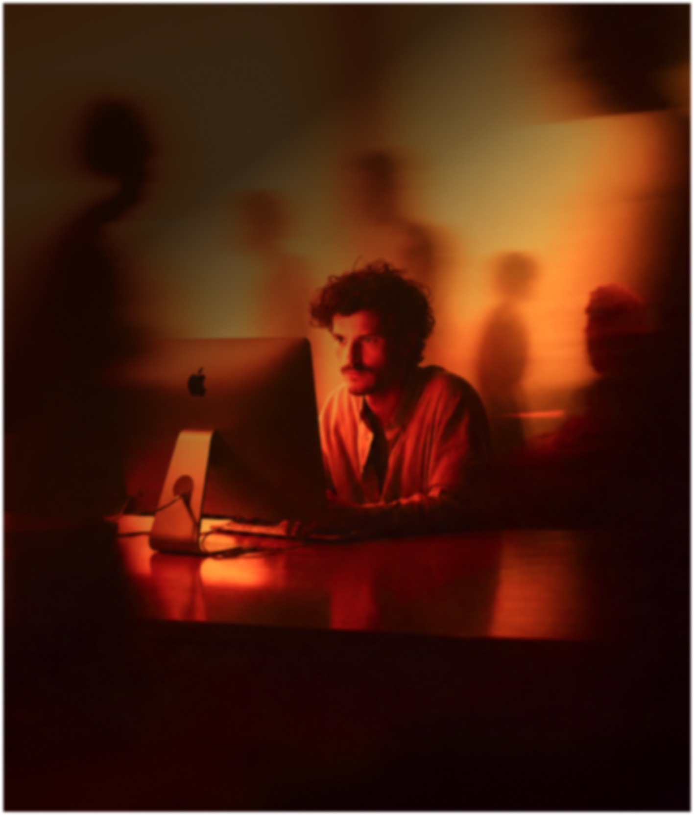 Man with curly hair sitting at a desk, focused on a large Apple iMac computer, with several blurred and shadowy figures in the background under warm orange lighting.