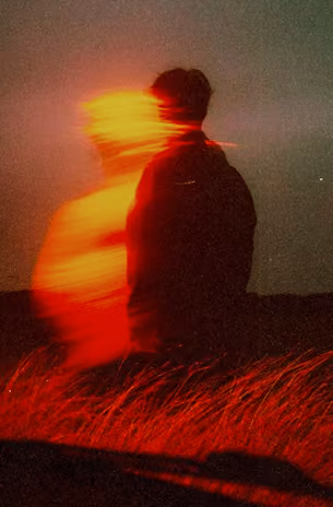 A silhouette of a person standing in a field with tall grass, illuminated by red light with an overlapping, blurred effect creating a ghostly second outline in yellow and red hues.
