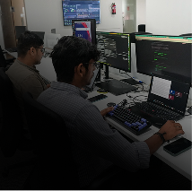Two men working at a desk with multiple monitors displaying code, with a laptop and other office equipment visible.
