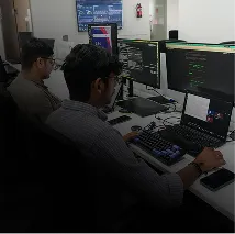 Two men working at a desk with multiple monitors displaying code, with a laptop and other office equipment visible.