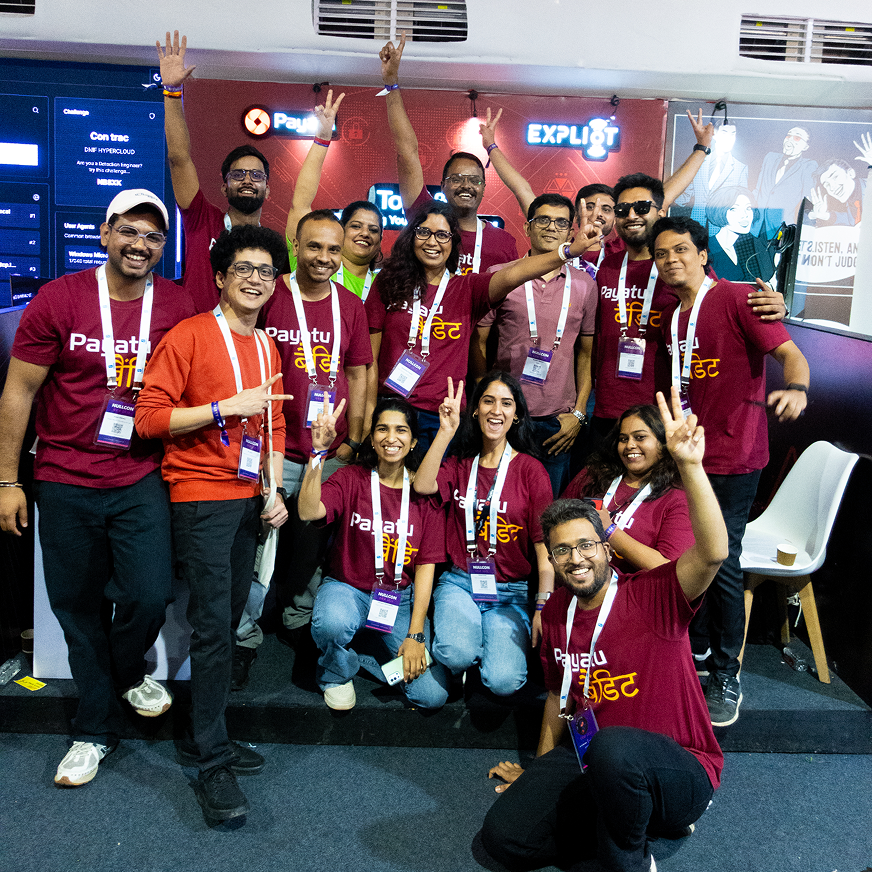 Group of smiling people wearing maroon Payatu t-shirts and event badges posing cheerfully with raised hands and peace signs at an indoor event booth.