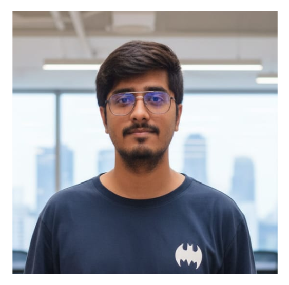 Young man with short dark hair, glasses with blue-tinted lenses, and a beard, wearing a dark shirt with a white Batman logo, standing indoors with blurred cityscape visible through large windows behind him.