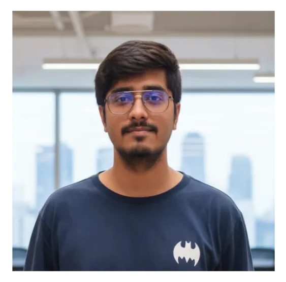 Young man with short dark hair, glasses with blue-tinted lenses, and a beard, wearing a dark shirt with a white Batman logo, standing indoors with blurred cityscape visible through large windows behind him.
