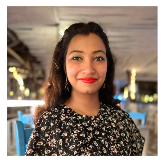 Smiling woman with long dark hair wearing red lipstick and a black floral top, with blurred lights and blue chairs in the background inside a softly lit indoor space.