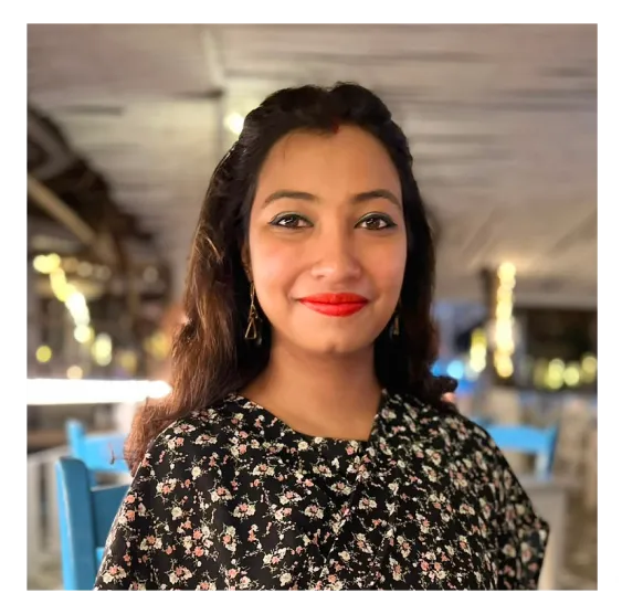 Smiling woman with long dark hair wearing red lipstick and a black floral top, with blurred lights and blue chairs in the background inside a softly lit indoor space.