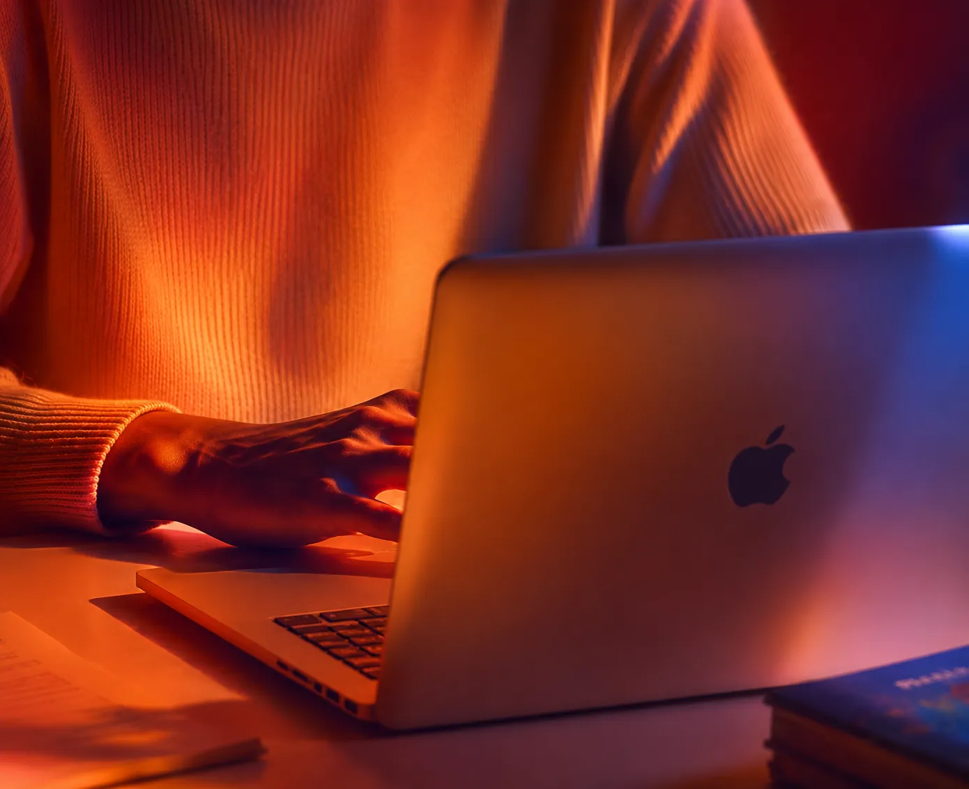 Person wearing a ribbed orange sweater typing on a silver Apple laptop on a desk lit with warm orange and blue lighting.