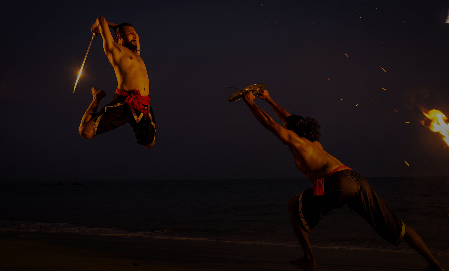 Two shirtless men on a beach at night engaged in a martial arts fight; one is airborne holding a glowing sword above his head ready to strike, the other is crouched holding a round shield defensively, with fire sparks visible on the right side.